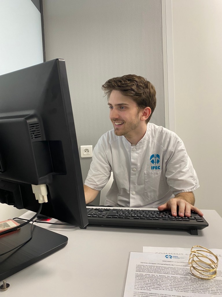 Mettre en avant le thérapeute Un chiropracteur à lyon souriant assis devant un ordinateur, vêtu d'une blouse blanche avec le logo IFEC, travaillant dans un bureau moderne.
